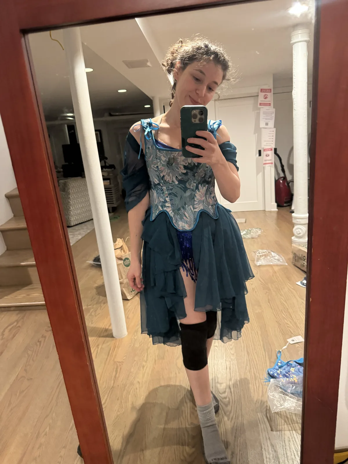 Mirror selfie wearing a blue corset with a layered dark teal skirt and one black knee pad.