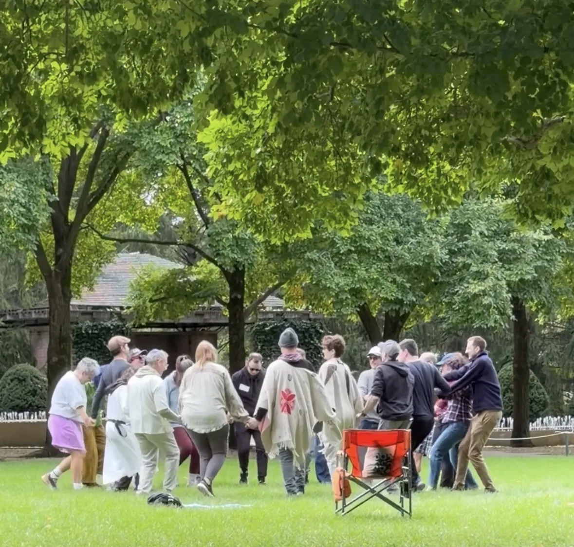 Event participants hold hands and move together in a circle on a grassy lawn beneath trees.